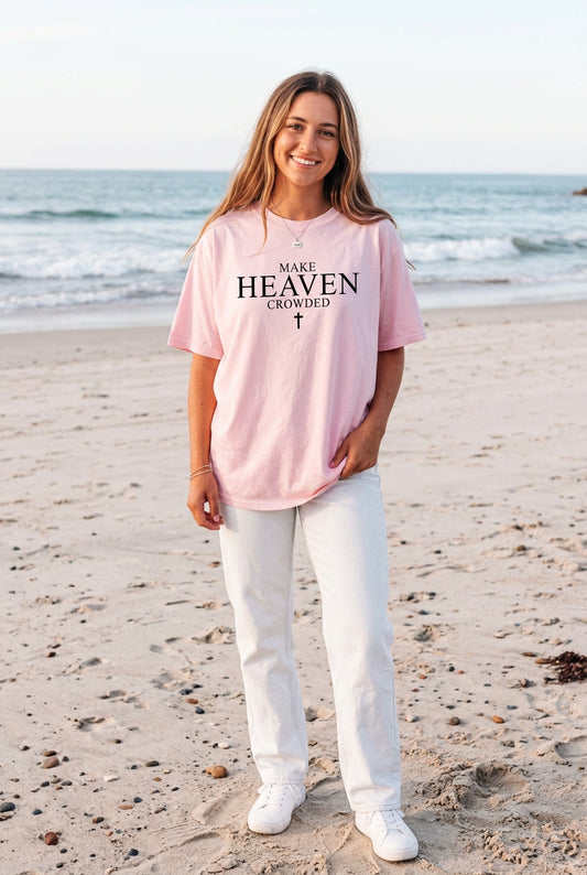 A woman wearing an oversized light pink t-shirt with the text 'MAKE HEAVEN CROWDED' and a cross printed in black, standing on a sandy beach with the ocean in the background.