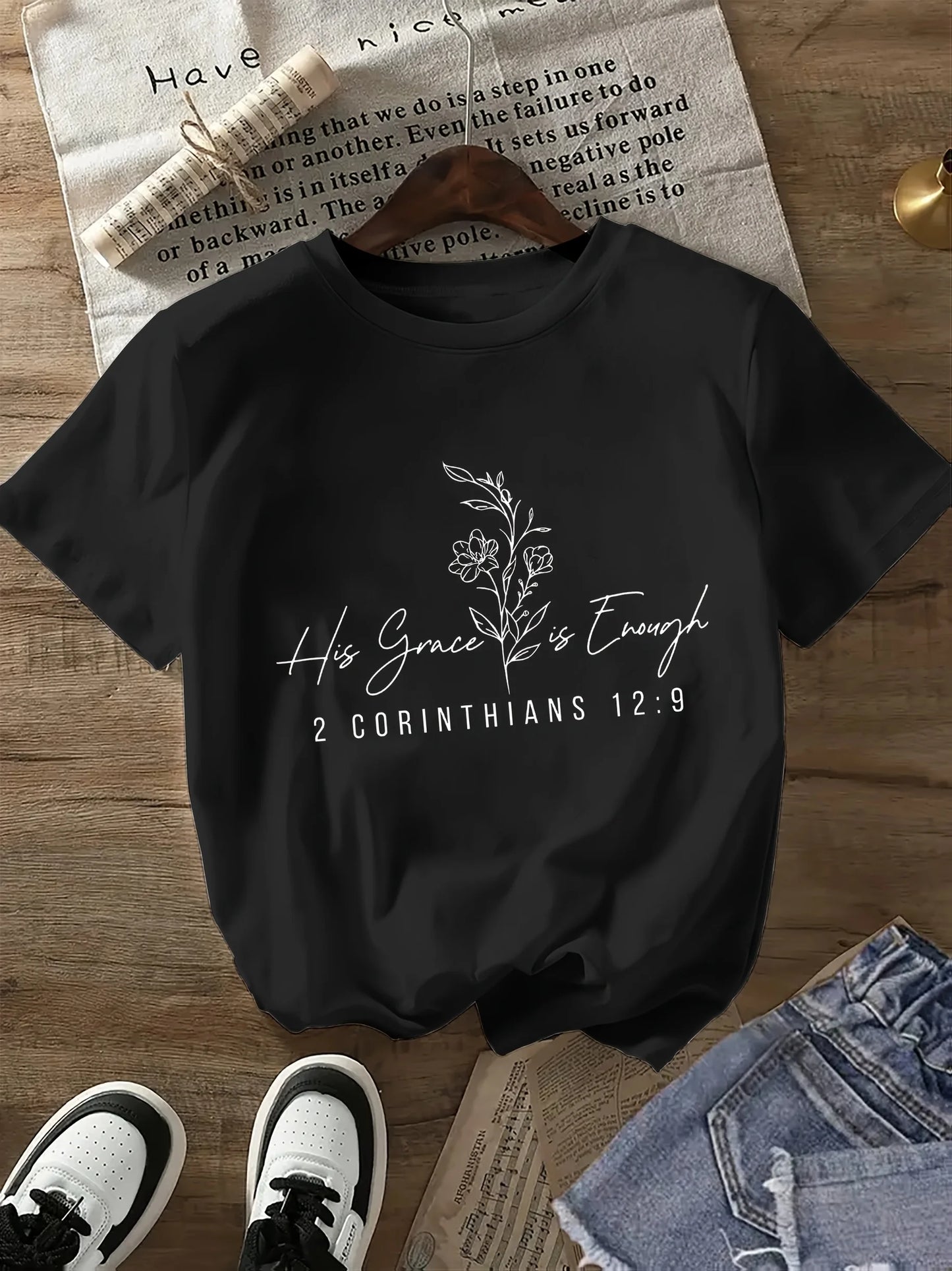 Black t-shirt with 'His Grace is Enough' text and floral design on a wooden surface with shoes and jeans.