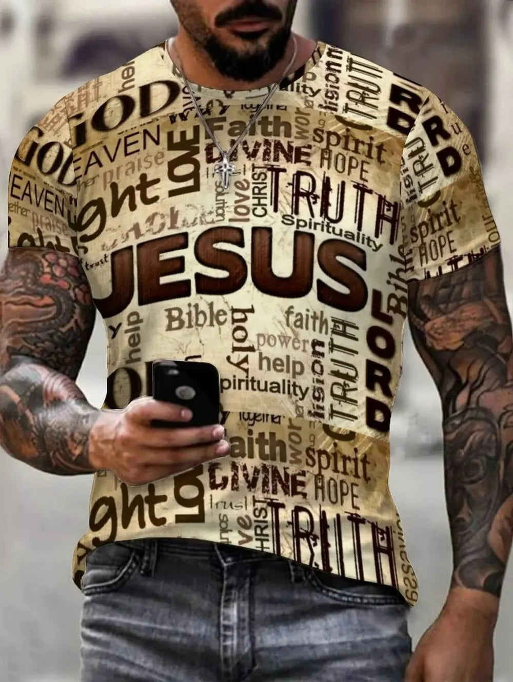 A man wearing a beige graphic t-shirt with a collage of religious words like 'GOD', 'JESUS', 'TRUTH', and 'SPIRIT' in dark brown and black text. He has tattooed arms and is holding a black smartphone.