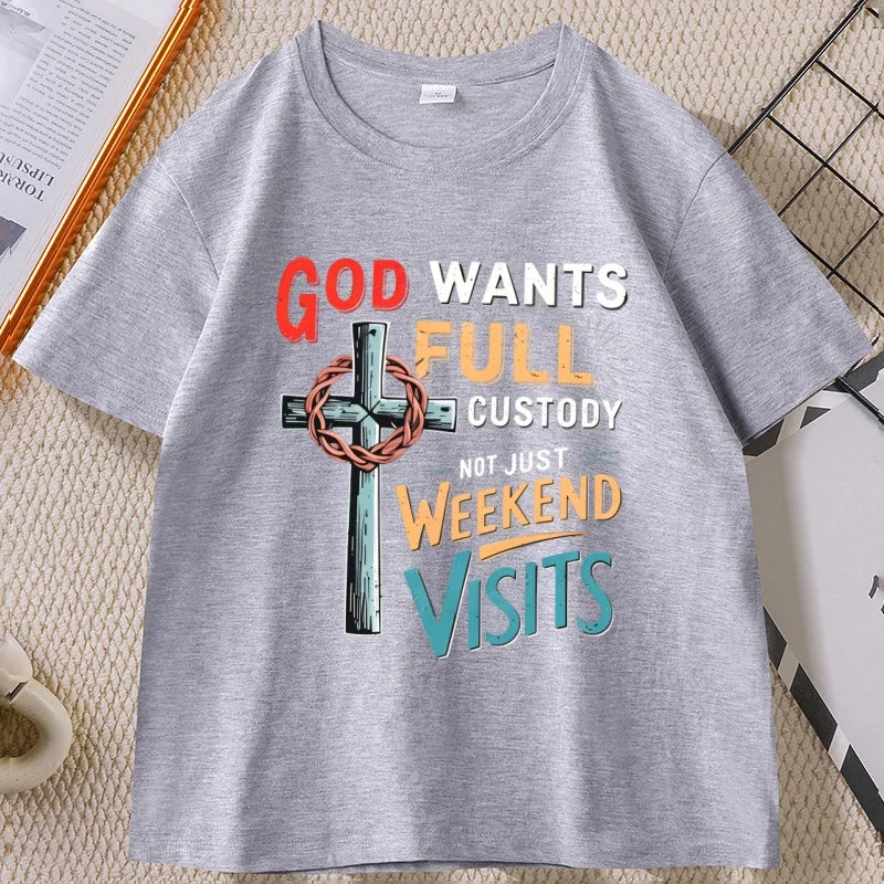 A heather gray short-sleeve t-shirt with a colorful graphic print featuring a cross and the text 'GOD WANTS FULL CUSTODY NOT JUST WEEKEND VISITS'. The t-shirt is laid flat on a textured beige woven mat with decorative props like a book and a wire basket.