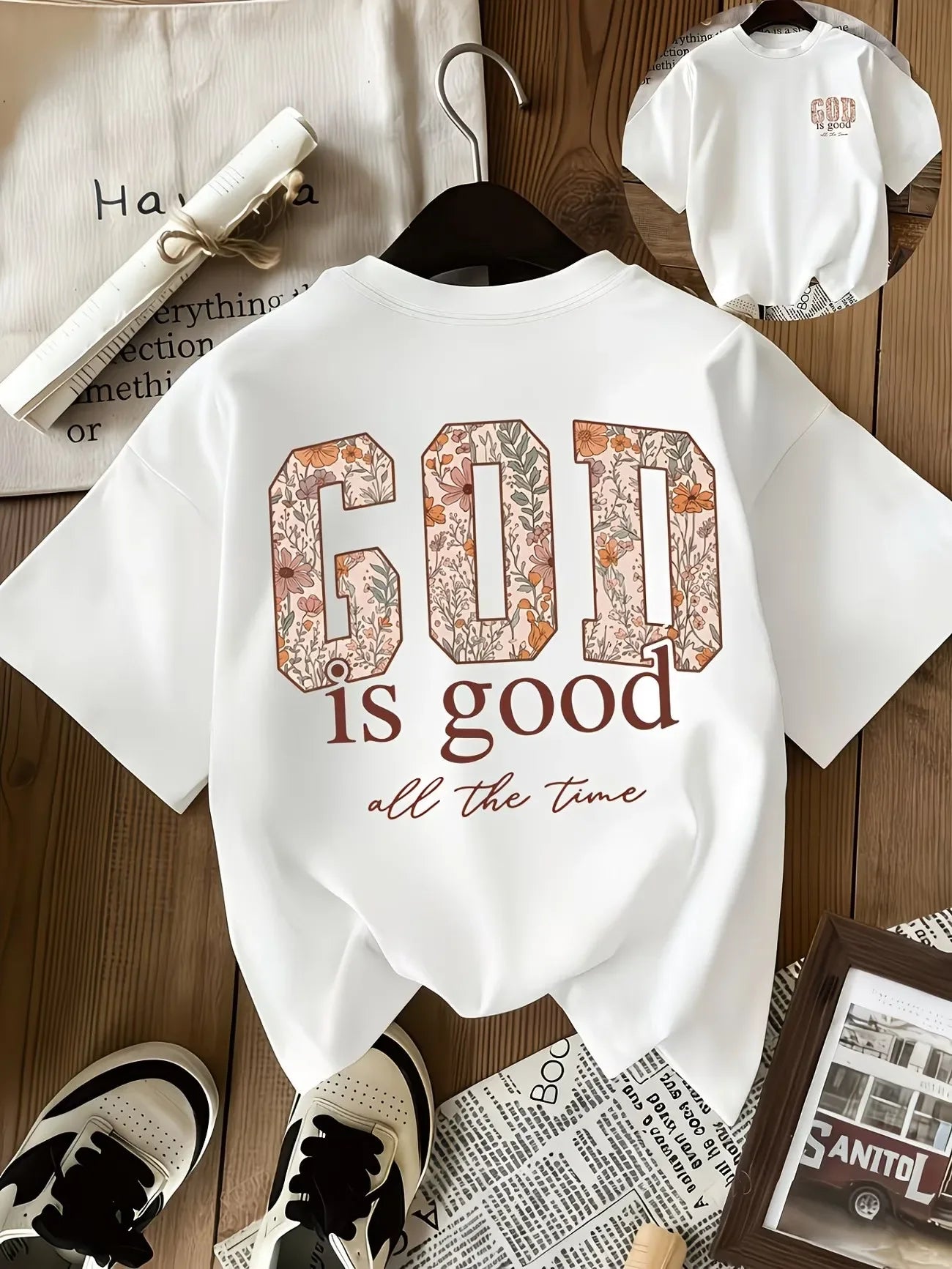 A white short-sleeve T-shirt hanging on a black hanger, featuring a large decorative 'GOD' graphic in a floral pattern above the words 'is good all the time' in brown script. The shirt is styled on a wooden surface with a small inset showing the front view, along with shoes, a rolled magazine, and framed print props.