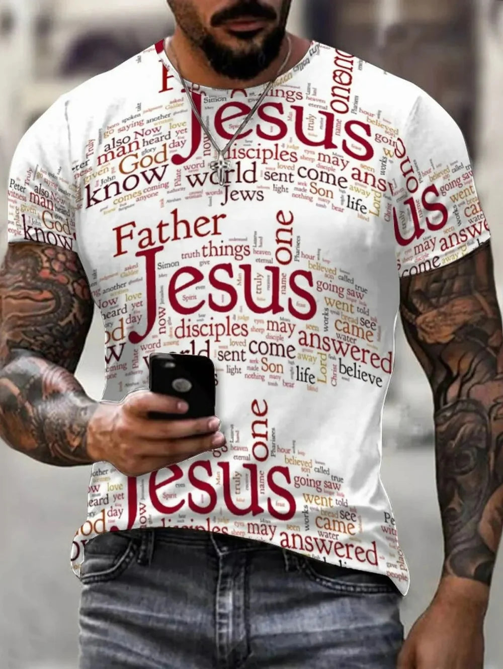A man wearing a white graphic t-shirt with a red and brown word cloud design featuring the word 'Jesus' and religious terms, holding a black smartphone.