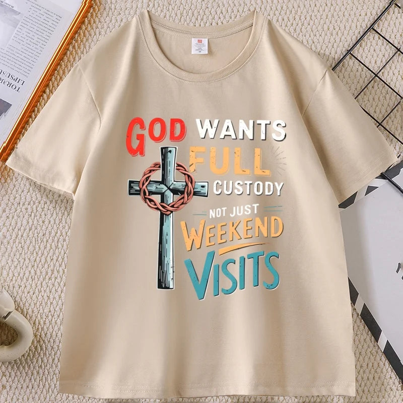 A beige short-sleeve t-shirt with a colorful graphic print featuring a cross with a crown of thorns and the text 'GOD WANTS FULL CUSTODY NOT JUST WEEKEND VISITS'. The t-shirt is laid flat on a textured beige surface with decorative props in the corners.