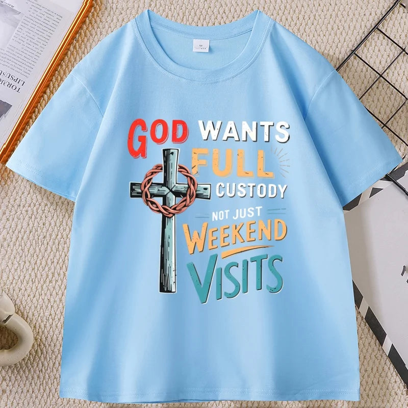 A light blue short-sleeve graphic T-shirt laid flat on a textured beige surface. The shirt features a large front print with the words 'GOD WANTS FULL CUSTODY NOT JUST WEEKEND VISITS' in red, white, yellow, and teal lettering, alongside a metallic cross with a wreath.