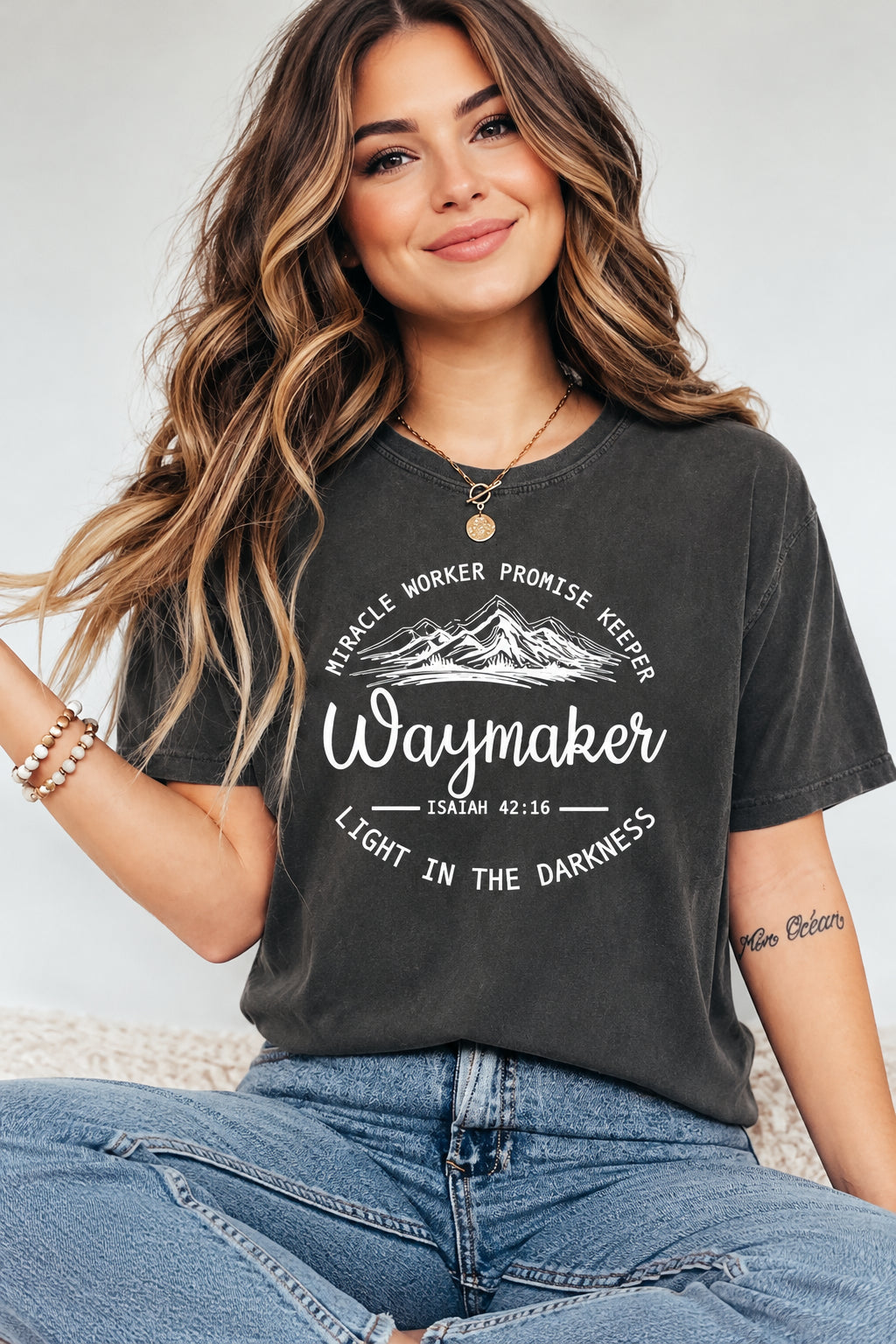 A woman wearing a dark charcoal gray washed graphic t-shirt with white text that reads 'Waymaker' and 'Miracle Worker Promise Keeper'.