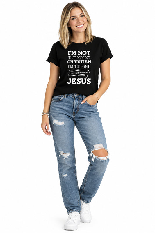 A woman wearing a black short-sleeve graphic T-shirt with white text that reads, 'I'M NOT THAT PERFECT CHRISTIAN I'M THE ONE THAT KNOWS I NEED JESUS,' paired with blue jeans against a plain white background.
