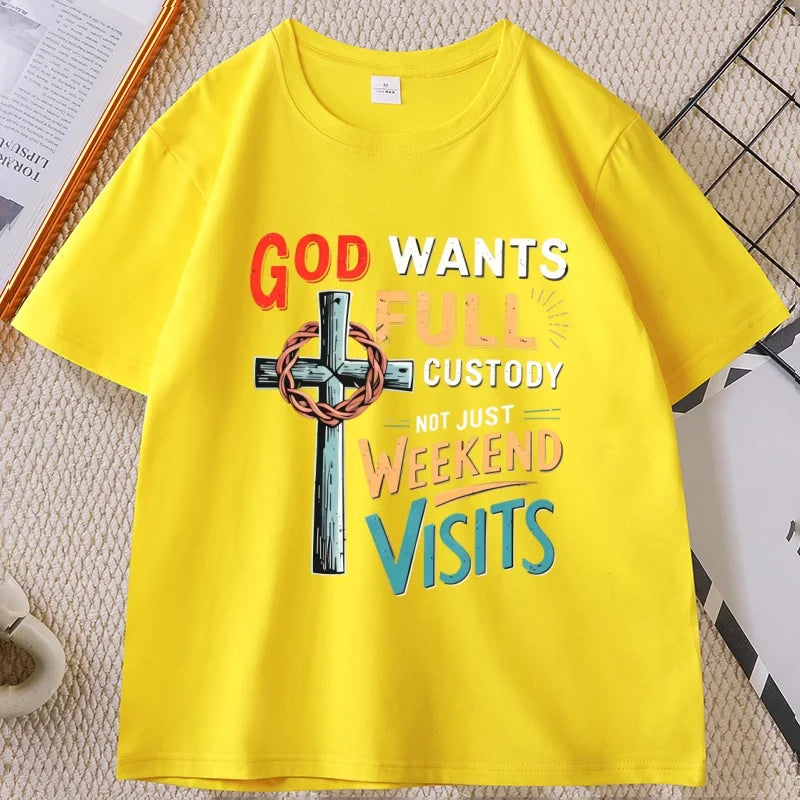 A bright yellow short-sleeve t-shirt with a colorful graphic print featuring a cross with a crown of thorns and the text 'GOD WANTS FULL CUSTODY NOT JUST WEEKEND VISITS'. The t-shirt is laid flat on a textured beige surface with decorative props.