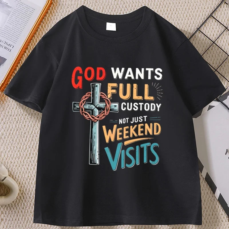 A black short-sleeve graphic T-shirt laid flat on a textured beige surface. The shirt features a colorful distressed typographic design with a cross and crown of thorns, reading 'GOD WANTS FULL CUSTODY NOT JUST WEEKEND VISITS' in red, white, yellow, and blue lettering.
