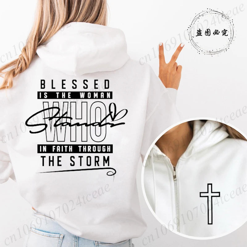 A woman shown from the back wearing a white hoodie with a black printed inspirational quote on the back. The text reads 'BLESSED IS THE WOMAN WHO STANDS IN FAITH THROUGH THE STORM' with a cursive 'STANDS' and a black cross symbol.