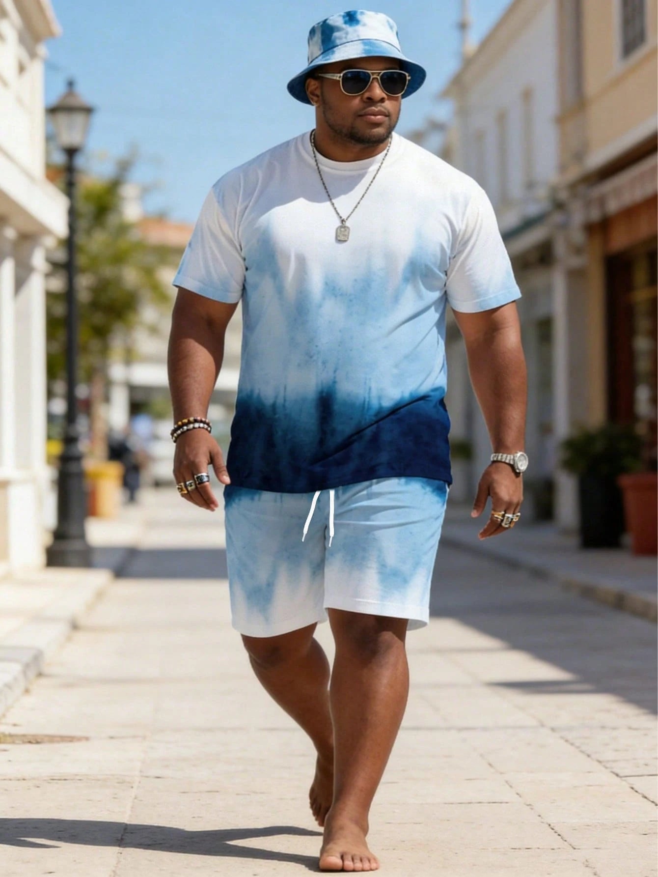 A man walking barefoot on a sunny outdoor street wearing a white and blue tie-dye t-shirt, matching tie-dye shorts, a blue bucket hat, and sunglasses.