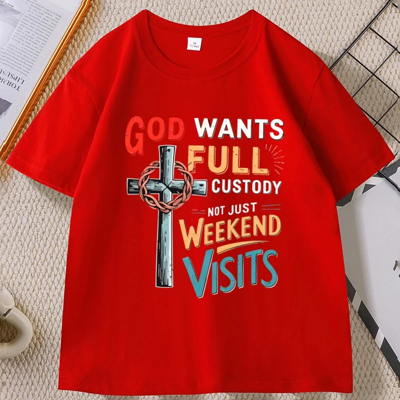A bright red short-sleeve t-shirt with a colorful graphic print featuring a cross and the text 'GOD WANTS FULL CUSTODY NOT JUST WEEKEND VISITS'. The shirt is laid flat on a textured beige surface with decorative props in the corners.