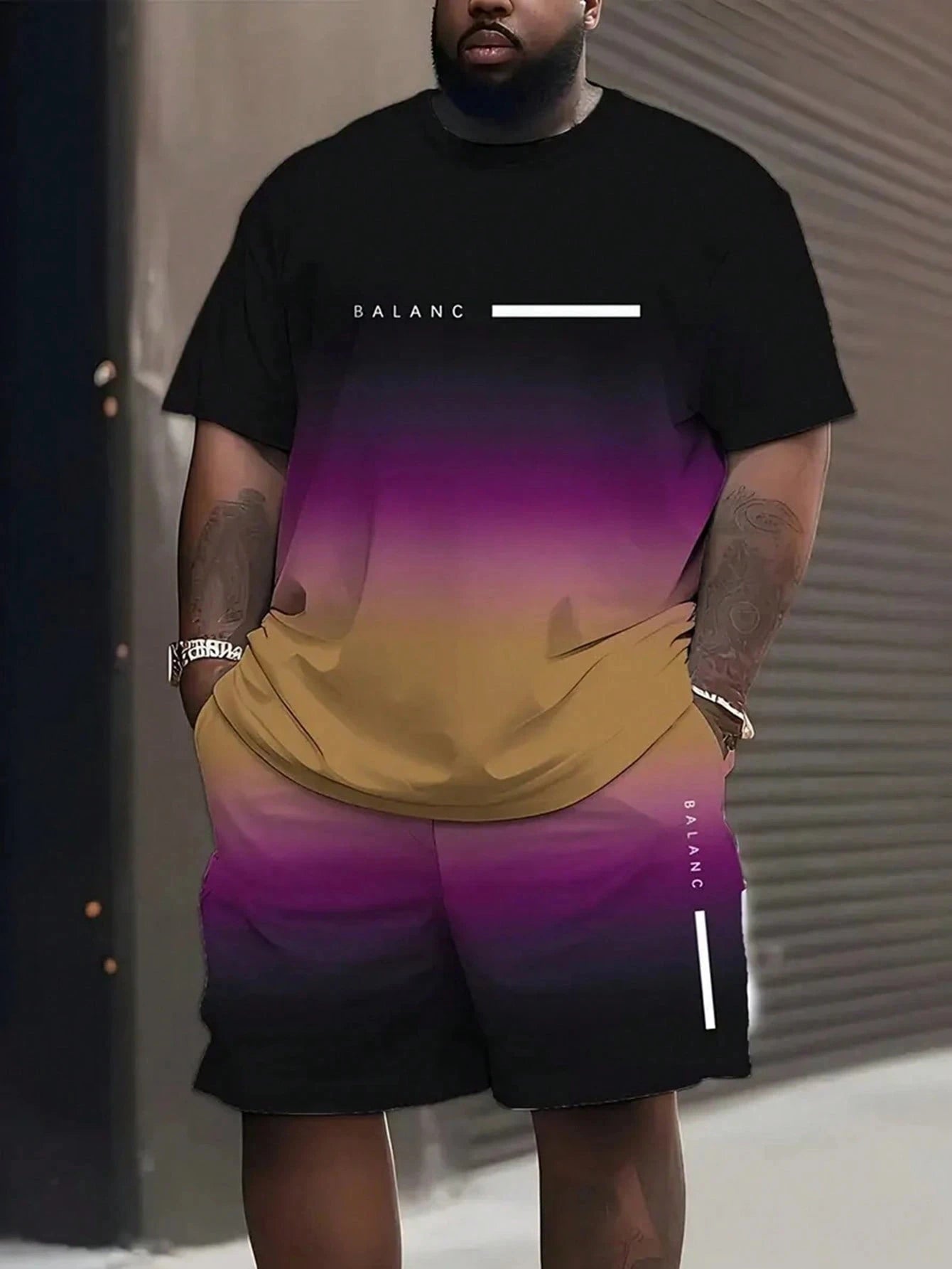 A man wearing a matching black t-shirt and shorts set with a purple-to-brown gradient design and white text accents, standing against a gray metal shutter background.