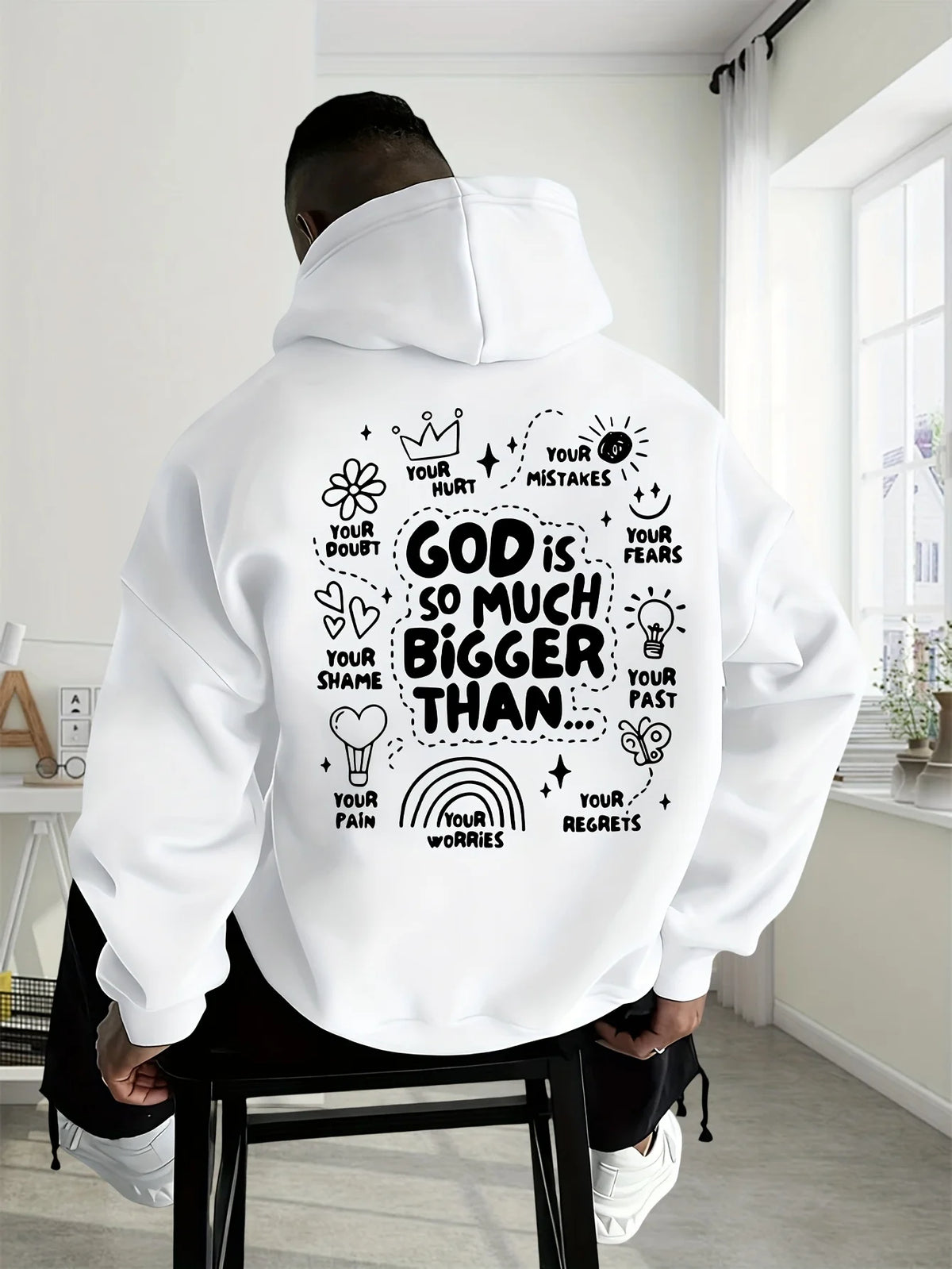 A man wearing a white hoodie from the back, seated on a black stool in a bright room. The hoodie features a large black hand-drawn style graphic with motivational text and doodles on the back.